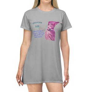 Puede incluir: Un vestido de camiseta gris con una ilustración de un gato rosa y el texto "GALATIAN 5:22" y una cita en texto azul. La cita dice "El fruto del Espíritu es amor, gozo, paz, paciencia, benignidad, bondad, fe, mansedumbre, templanza."