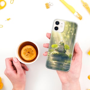 May include: A clear phone case with a green frog wearing a gold crown sitting on a lily pad in a pond. The background is a blurry green and brown.