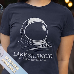 Puede incluir: Camiseta azul marino con una ilustración blanca de un casco de astronauta emergiendo del agua. El texto "LAKE SILENCIO" y las coordenadas están impresas debajo. Disponible en 12 colores.