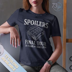 Puede incluir: Camiseta azul marino con las palabras blancas "SPOILERS FINAL TOUR 24 YEARS AT DARIILIUM". El gráfico incluye una imagen similar a un libro. La camiseta está disponible en 12 colores.