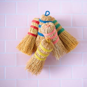 May include: Five small, natural fiber brooms with colorful yarn handles. The brooms are tied together and hanging against a pink tiled wall.