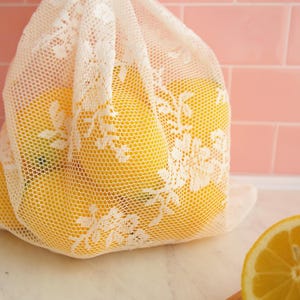 May include: A white lace mesh produce bag filled with yellow lemons. The bag is sitting on a white countertop.
