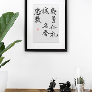 May include: A framed artwork with black calligraphy on a white background. Below, a black miniature scooter and a glass terrarium with a small plant sit on a wooden surface. A large green plant in a white pot is on the left.