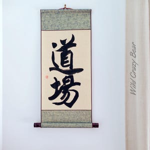 May include: A traditional Japanese scroll with black calligraphy on a cream background. The scroll is framed with a green and gray patterned border and a dark brown wooden rod at the bottom. The text is in Japanese. A bouquet of dried flowers is in the foreground.