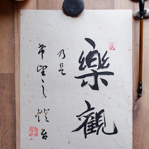 May include: A calligraphy artwork on textured paper, featuring black ink characters and red stamps. The artwork is centered on a wooden surface, with a ceramic cup and ink brush visible in the background. The calligraphy is in a traditional East Asian style.