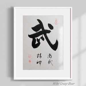 May include: A framed artwork featuring Japanese calligraphy in black ink on textured paper. The main character is bold, with additional characters and red stamps. The frame is white, and the artwork is against a light gray wall.