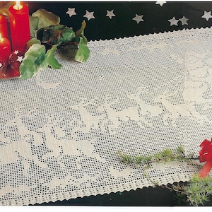Op de afbeelding: Witte gehaakte tafelloper met een kerstthema met rendieren en een kerstboom. De loper ligt op een donker oppervlak met een groene plant en rode ornamenten.