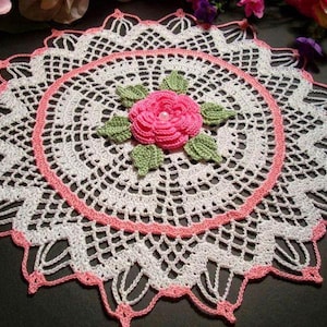 May include: A white crochet doily with a pink crocheted flower in the center. The doily has a scalloped edge and is made with delicate stitches.
