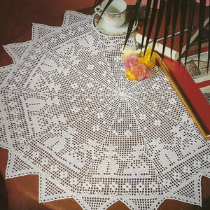 May include: A white, star-shaped, crocheted doily. The intricate lace pattern features geometric and floral designs. A teacup and saucer, along with a small bouquet of flowers, are placed on the doily. The doily is on a brown surface.