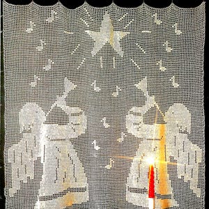 May include: A white, crocheted curtain featuring a star and angel design. The curtain is hung with gold-colored rings. A lit red candle is in the foreground. The curtain's design includes musical notes and angel figures, creating a festive, holiday theme.