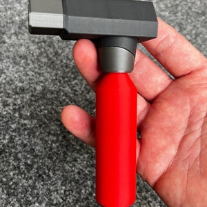 May include: A red and black tool with a cylindrical red base and a rectangular black top. The tool is held in a hand against a gray background. The tool appears to be a specialized device.