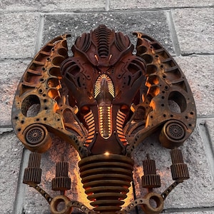 May include: A rusty metal wall sculpture in the shape of a stylized alien head with glowing orange lights. The sculpture is made of various metal parts and has a steampunk aesthetic.