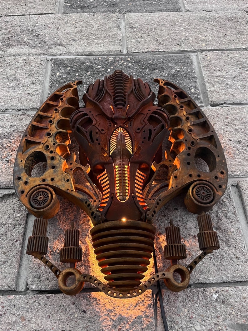 May include: A rusty metal wall sculpture in the shape of a stylized alien head with glowing orange lights. The sculpture is made of various metal parts and has a steampunk aesthetic.