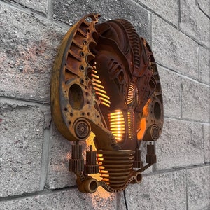 May include: A wall-mounted light fixture made from repurposed metal parts. The light fixture is shaped like a face with a rusty brown finish and a glowing orange light.