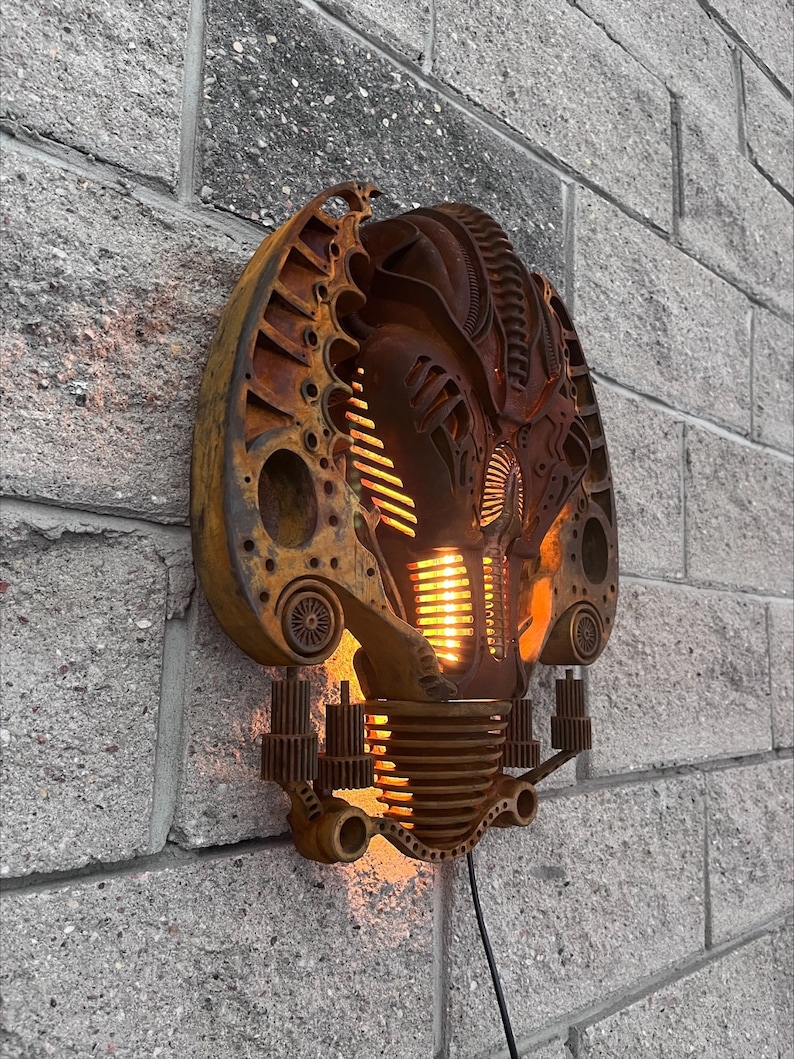 May include: A wall-mounted light fixture made from repurposed metal parts. The light fixture is shaped like a face with a rusty brown finish and a glowing orange light.