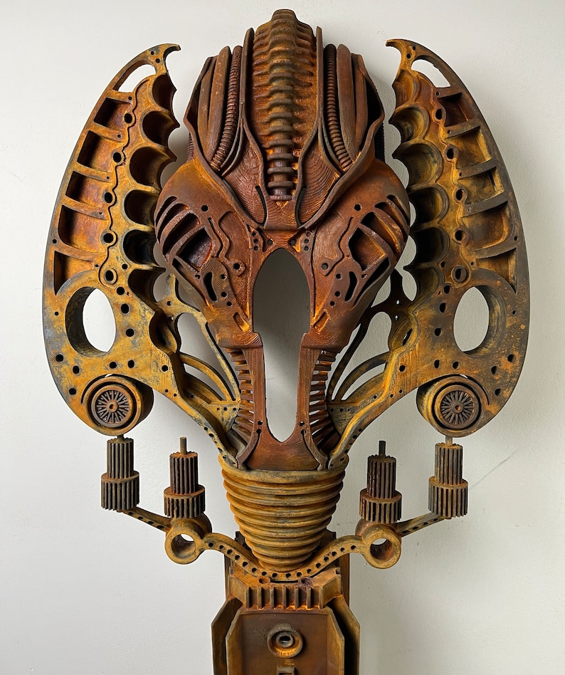 May include: A rusty metal sculpture of a stylized alien head with intricate details and a textured surface. The sculpture is mounted on a base with gears and other mechanical elements.