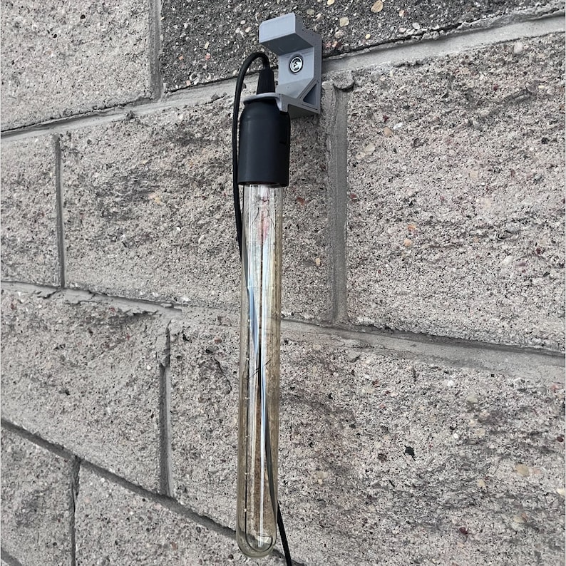 May include: A clear glass tube light bulb with a black cord and a gray wall mount. The light bulb is hanging from the wall mount.