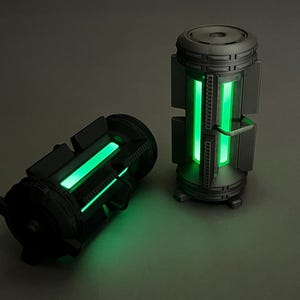 May include: Two dark gray cylindrical containers with glowing green lights. One container is upright, and the other is on its side. The containers have a futuristic design, with visible mechanical details and a bright green glow.
