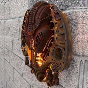 May include: A rusty metal wall sculpture in the shape of a face with intricate details and a glowing light inside. The sculpture is mounted on a brick wall.