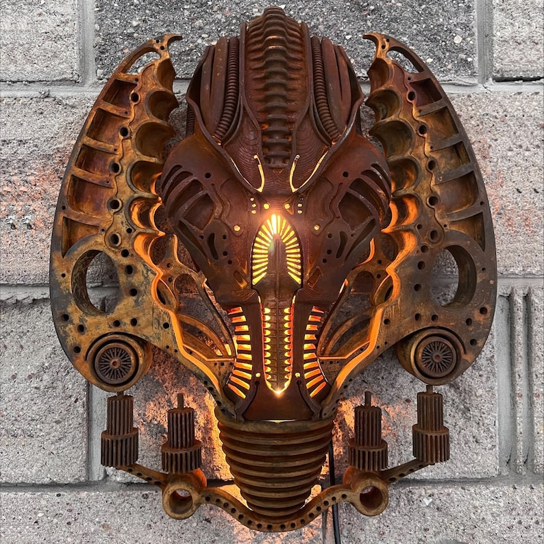 May include: A rusty metal wall sculpture in the shape of a face with intricate details and a glowing orange light in the center.