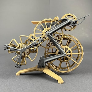 Puede incluir: Un modelo de mecanismo de reloj con engranajes de color dorado y un marco estructural gris oscuro. El conjunto incluye un volante central con un resorte en espiral, ruedas dentadas interconectadas y una base de trípode, expuesto como una escultura cinética.