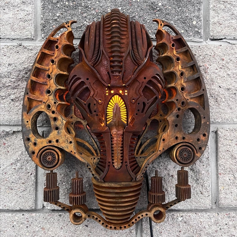May include: A rusty metal wall sculpture in the shape of a face with intricate details and glowing lights. The sculpture is mounted on a brick wall.