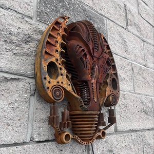 May include: A rusty metal wall sculpture in the shape of a face with intricate details and gears. The sculpture is mounted on a brick wall.