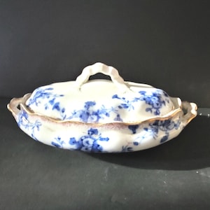 May include: A white porcelain serving dish with a lid, adorned with blue floral patterns. The dish has a scalloped edge with a gold trim and a handle on the lid. The dish is oval-shaped.