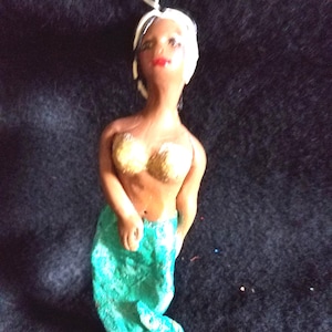 May include: A handmade mermaid figurine with a brown body, gold-colored top, and a turquoise tail. The mermaid has white hair and red lips. The figurine is suspended by a white string, suggesting it's an ornament.
