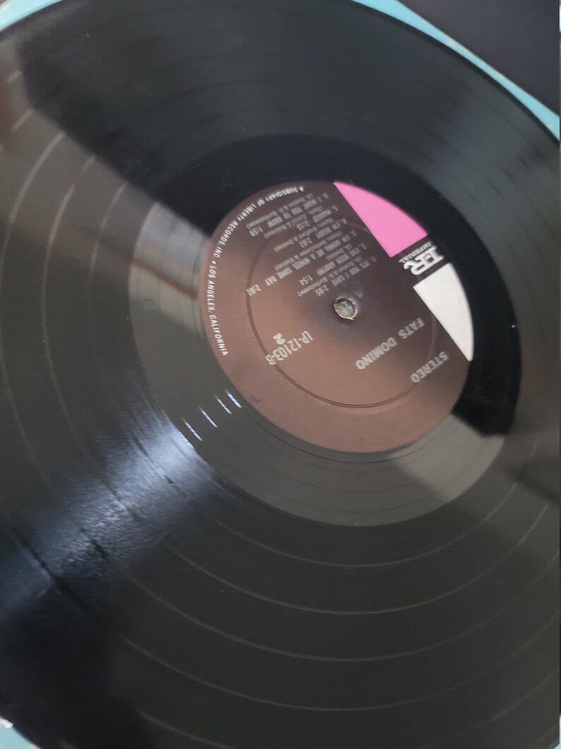 Puede incluir: Un disco de vinilo negro con el texto "Fats Domino" y "Stereo". La etiqueta del disco es marr&oacute;n y rosa, con t&iacute;tulos de canciones y duraciones. Un art&iacute;culo de m&uacute;sica vintage.