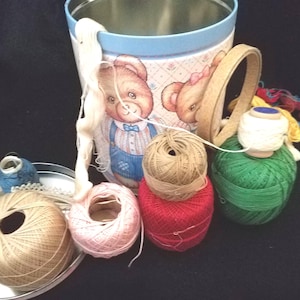 May include: A selection of colourful thread and yarn spools, including white, pink, red, green, and tan, are arranged near a decorative tin with a teddy bear design. A metal tray holds additional thread spools. The scene suggests a crafting or sewing theme.