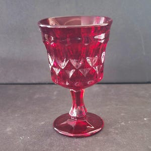 Könnte beinhalten: Ein rubinroter Glasbecher mit einem Diamant- und Herzmuster. Stiel und Fuß sind ebenfalls rot. Der Becher wird wahrscheinlich zum Servieren von Wein oder anderen Getränken verwendet. Das Glas hat einen Vintage-Look.