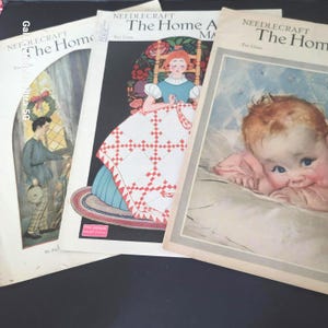 Könnte beinhalten: Ein Stapel alter Needlecraft-Magazine. Die Cover zeigen Illustrationen einer Frau, die eine Steppdecke näht, eines Babys und eines Mannes, der aus einem Fenster schaut. Die Magazine tragen den Titel "The Home".