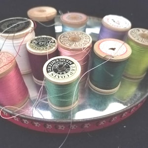 Pode incluir: Uma coleção de bobinas de linha vintage em várias cores, incluindo rosa, verde e roxo, dispostas numa bandeja de metal decorativa com uma borda floral vermelha. As bobinas têm núcleos de madeira e são rotuladas com texto.