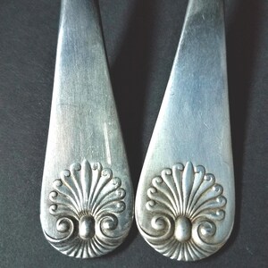 May include: Two silver serving spoons with a decorative, symmetrical design at the base. The design features a central oval shape with radiating leaf-like elements and scrolled details. The spoons are set against a dark background.