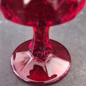 Op de afbeelding: Close-up van een robijnrode glazen beker. De steel en voet zijn zichtbaar en tonen het ingewikkelde geslepen glasontwerp. De kleur van de beker is diep, rijk rood, met een glanzende afwerking. De voet is rond en de steel is kort.