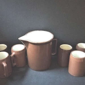 May include: A brown ceramic pitcher with a white interior and a handle, surrounded by six matching mugs. The mugs and pitcher have a similar design, with a brown exterior and white interior. The set is likely for serving beverages.