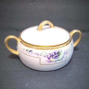May include: A white porcelain sugar bowl with a lid and two handles, accented with gold trim. The bowl features a floral design with purple and green flowers. The lid has a gold handle.