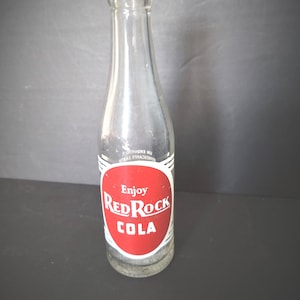 May include: A clear glass Red Rock Cola bottle with a red and white label. The label reads "Enjoy Red Rock COLA" in white text on a red oval. The bottle has a narrow neck and a rounded base.