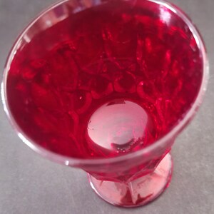 Op de afbeelding: Een helder glas met voet, gevuld met een helderrode vloeistof. Het glas heeft een facetontwerp en een ronde basis. De vloeistof lijkt kleine belletjes te hebben, wat wijst op een koolzuurhoudende drank of cocktail. Het glas staat op een grijze achtergrond.