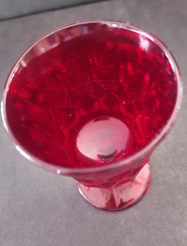 Op de afbeelding: Een helder glas met voet, gevuld met een helderrode vloeistof. Het glas heeft een facetontwerp en een ronde basis. De vloeistof lijkt kleine belletjes te hebben, wat wijst op een koolzuurhoudende drank of cocktail. Het glas staat op een grijze achtergrond.