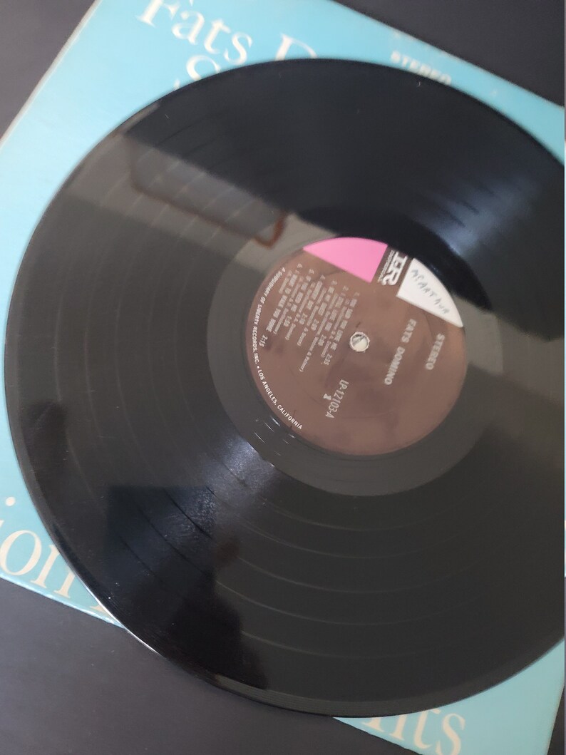 Puede incluir: Un disco de vinilo negro de Fats Domino, etiquetado como "STEREO". El disco es parcialmente visible, descansando sobre una portada de &aacute;lbum azul claro. La portada del &aacute;lbum tiene el texto "Fats Domino" y otros textos.