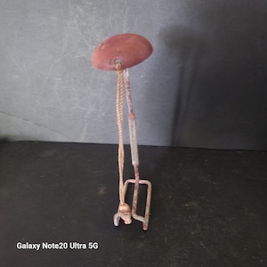 May include: A vintage metal tool with a red, rounded top and a spring mechanism. A rope hangs from the top, connected to a small, hook-shaped piece at the bottom. The tool has a weathered, rustic appearance.