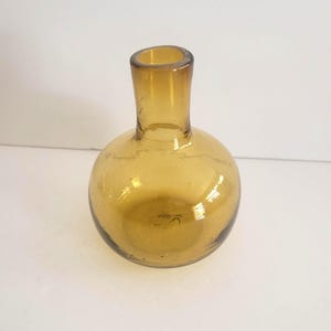 Peut inclure: Petite bouteille en verre ambré, corps arrondi et col étroit. La bouteille a une esthétique vintage, avec de légères imperfections dans le verre. Convient à des fins décoratives.