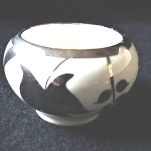 May include: A small, round, white ceramic bowl with a black floral design. The rim is silver. The bowl is sitting on a dark surface. The bowl is likely for decorative purposes.