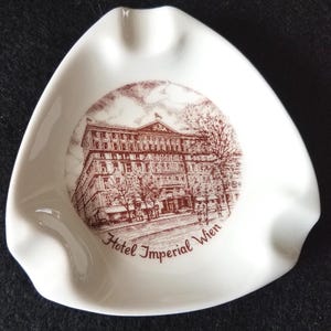 May include: White ceramic ashtray with a triangular shape. The center features a brown illustration of a building with the text "Hotel Imperial Wien". The ashtray has three curved indentations for resting cigarettes.