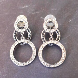 May include: A pair of silver-toned dangle earrings. Each earring features three connected circles, with the largest at the bottom. The circles are embellished with clear and black rhinestones. The earrings have a post backing.