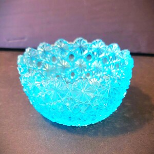 Könnte beinhalten: Eine kleine, runde, türkisfarbene Glasschale. Die Schale hat eine strukturierte Oberfläche mit einem sich wiederholenden Sternmuster. Der Rand der Schale ist gewellt. Die Schale steht auf einer dunklen Oberfläche.