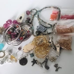 Pode incluir: Uma coleção de materiais para fazer joias, incluindo pulseiras douradas, contas, berloques e fechos. Há também vários sacos pequenos de contas em várias cores, juntamente com alguns círculos de madeira e um alfinete com a palavra "Inspire".
