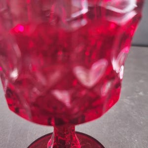 Op de afbeelding: Een robijnrode glazen beker met een hartvormig patroon. De steel en voet zijn ook rood en het glas heeft een textuur. De beker is waarschijnlijk bedoeld voor het serveren van dranken.